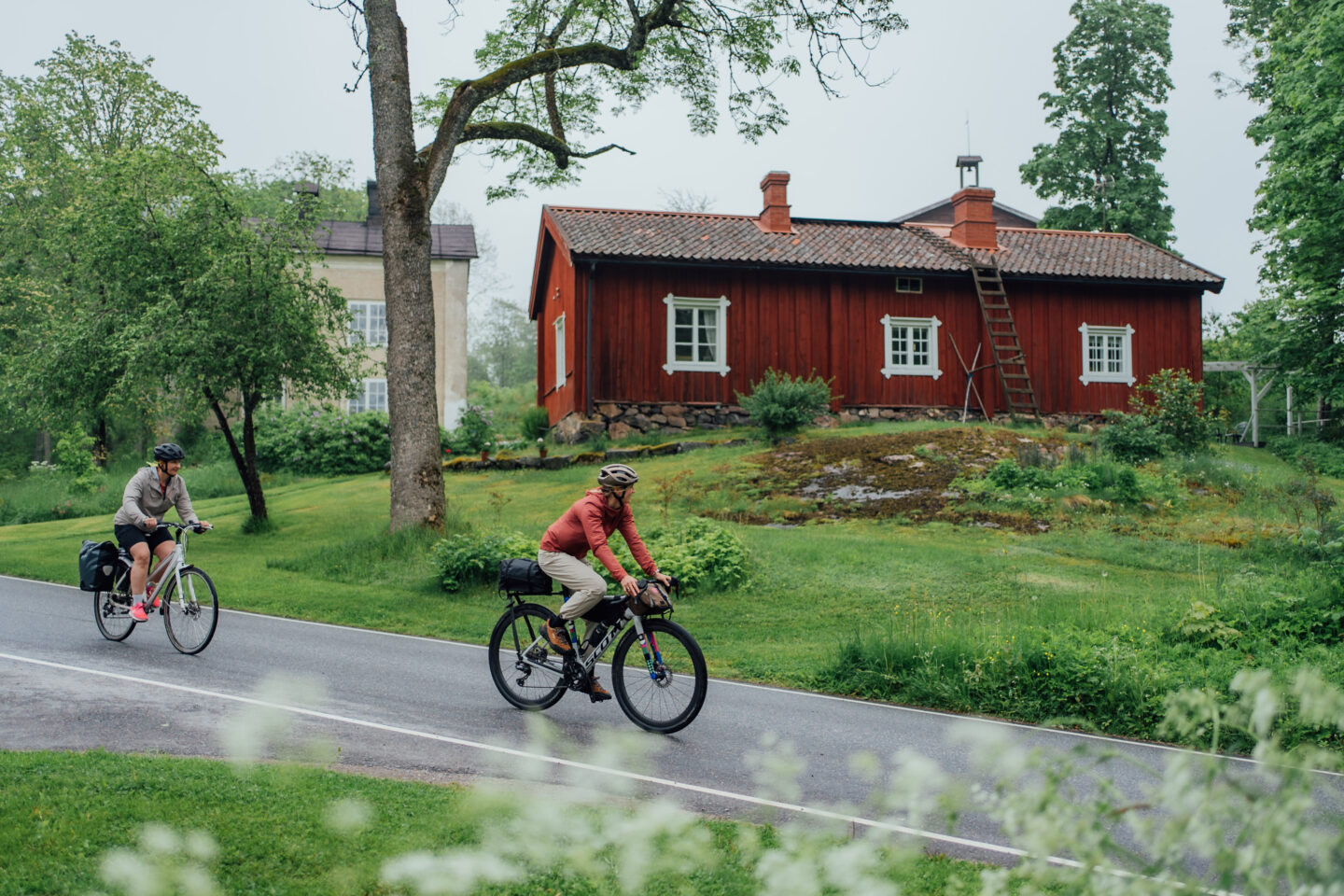 Two cyclists riding a paved road past some wooden country houses
