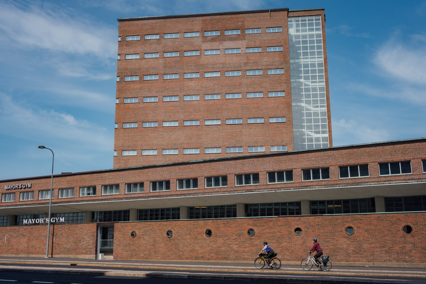 Two cyclists riding a cycling road besides a big red brick building