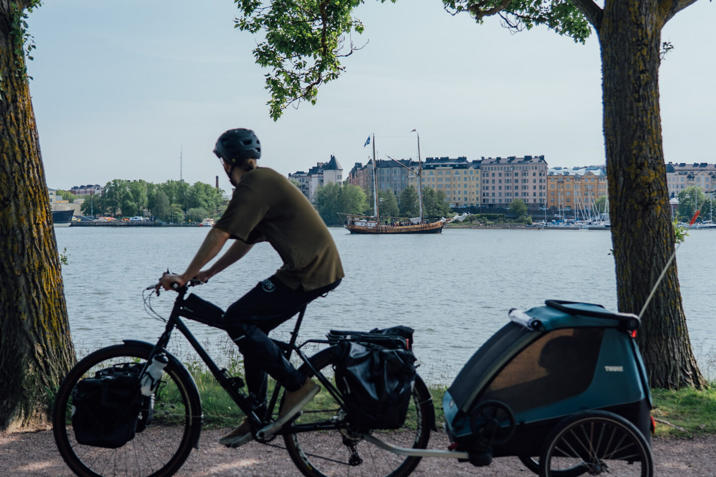 A cyclist riding a gravel road next to waterfront, with a baby trailer, looking at a boat in front of a city view