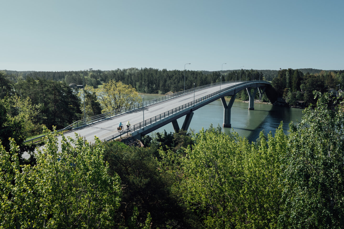 An air picture of two cyclists riding over a big river bridge, with a foresty view