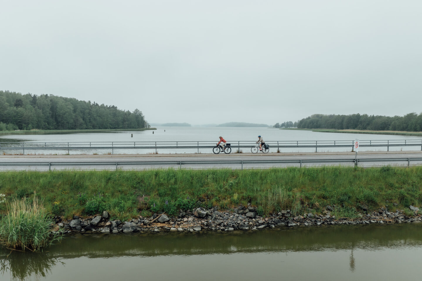Two cyclists riding a paved road over a bank in a lake, on a cloudy day