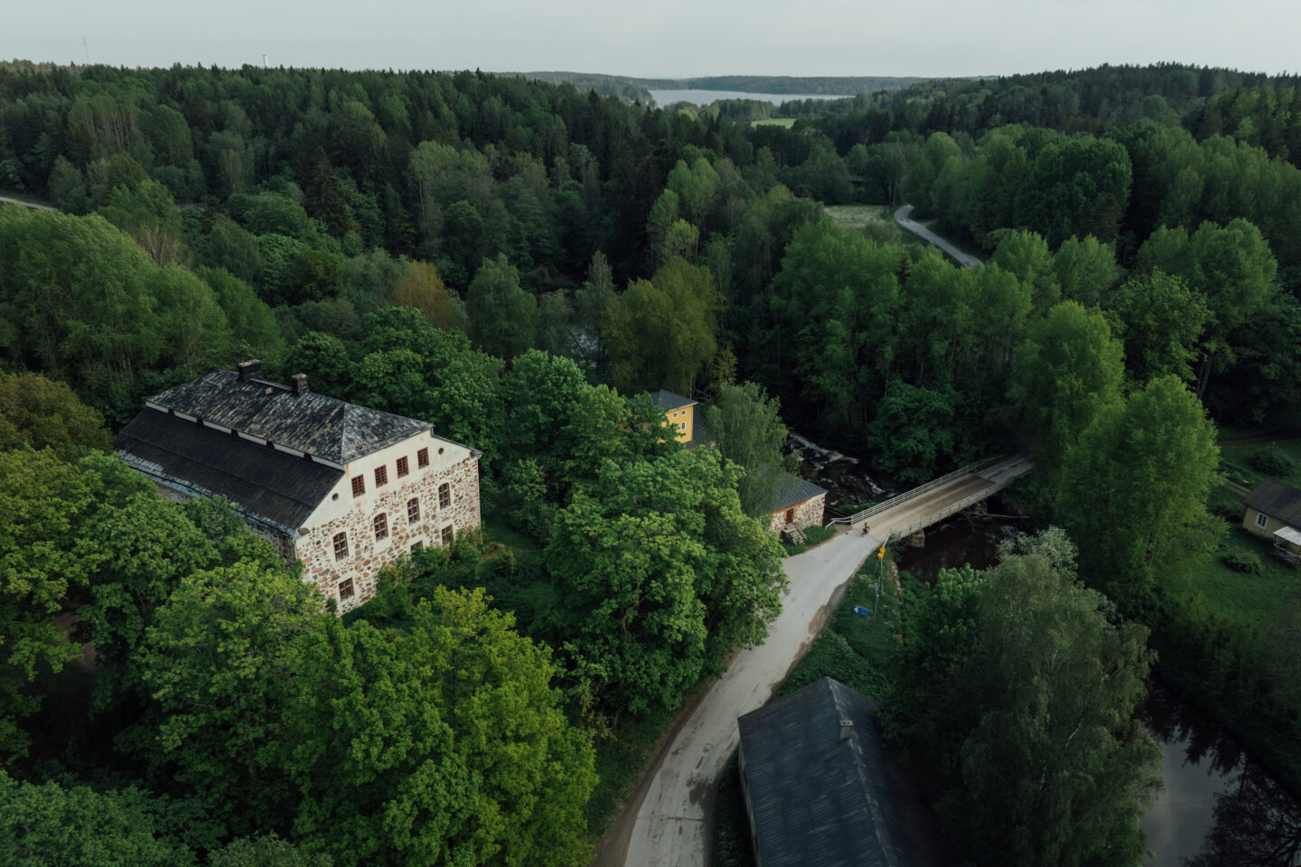An air picture of a cyclist riding a paved road that goes through a forest, over a river bridge and past some old stone buildings